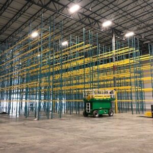Interior of a large warehouse featuring tall, empty metal pallet rack shelving units in green and yellow. The pallet rack shelves are arranged in rows, creating a spacious storage area. There are two green scissor lifts parked in front of the pallet rack shelves, indicating ongoing or recent installation work. The high ceiling with industrial lighting provides bright illumination throughout the space. The concrete floor is clean and the overall environment appears organized and well-maintained.