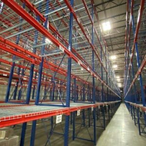 Interior of a large warehouse with long rows of tall, blue and red metal pallet rack shelving units. The pallet rack shelves are empty, with wire decking in place to hold items. The pallet rack shelves extend far into the distance, creating a sense of depth and vast storage capacity. The high ceiling with bright industrial lighting provides ample illumination throughout the space. The clean concrete floor adds to the orderly and spacious environment.