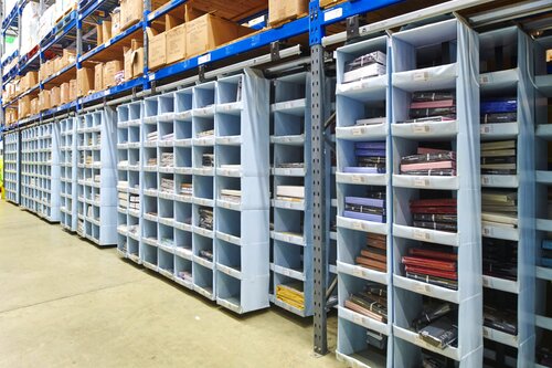 A well-organized Storeganizer warehouse section features multiple shelving units filled with light blue storage compartments. Each compartment holds neatly stacked items, such as folded fabrics or garments, sorted by color and type. The shelving units have grey metal frames and blue horizontal supports, providing a sturdy structure for the compartments. Above the compartments, larger boxes are stored on higher shelves, adding to the efficient use of vertical space. The concrete floor is clean and clear, allowing easy access to the items. The overall scene reflects a highly organized storage system designed for easy identification and retrieval of inventory.