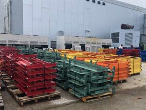 The image shows an outdoor storage area with stacks of used metal shelving components organized on wooden pallets. The components are painted in various colors, including red, green, yellow, and orange, and are neatly stacked and banded together for stability. The pallets are arranged in rows, indicating an organized inventory system. In the background, there are large industrial units and two people engaged in some activity. The setting suggests that these shelving components are ready for sale or reuse, awaiting transportation or assembly in a different location. The overall scene depicts a well-maintained and orderly outdoor storage area for industrial materials.