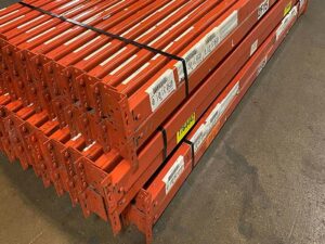 The image shows a stack of used orange metal rails, neatly banded together and ready for sale. The rails have several labels and barcodes attached to them, indicating their specifications and inventory information. The ends of the rails are designed with multiple holes for adjustable assembly in a shelving or racking system. The rails are stacked on a concrete floor, with visible wear and markings that suggest previous use. Despite being used, the rails appear to be in good condition and are organized efficiently for transport or storage.