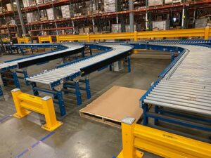 A warehouse interior features a used powered conveyor system with metal rollers and sturdy blue frames. The conveyor has a curved section, allowing it to navigate turns efficiently. Bright yellow safety barriers are installed around the conveyor to protect equipment and personnel. In the foreground, a wooden pallet with a large sheet of cardboard is placed on the floor. The background shows rows of tall shelving units filled with various boxes and inventory, indicating a well-organized storage area. The clean, polished concrete floor and the systematic arrangement of the conveyor system highlight the facility's focus on efficiency and safety in material handling operations.