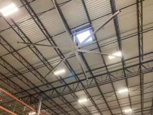The image shows a large ceiling fan installed in a warehouse. The fan has multiple long blades and is suspended from the high ceiling, which is constructed with metal beams and panels. The fan is designed to provide air circulation in the expansive warehouse space. The ceiling also features bright industrial lights, and there is a skylight visible near the fan, allowing natural light to enter. The overall setup indicates an effort to maintain a well-ventilated and well-lit environment in the warehouse.