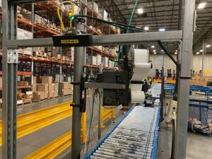 A warehouse setting features a used Cognex automated labeling machine mounted on a metal frame above a conveyor system with metal rollers and a blue frame. The machine is equipped with rolls of labels and various electronic components for automated operation. In the background, tall shelving units filled with boxes and inventory are visible, along with bright yellow safety barriers. The warehouse has a high ceiling with industrial lighting, providing ample illumination. A worker wearing a high-visibility vest can be seen in the distance, indicating an active and organized workspace focused on efficient material handling and processing