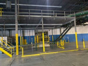 The image shows a used warehouse mezzanine structure. The mezzanine is elevated with a sturdy metal frame, supported by vertical columns with yellow safety padding at their bases. There are stairs with yellow railings leading up to the mezzanine level, which appears to have metal grating for the floor. Below the mezzanine, there are several yellow safety bollards and a conveyor system, indicating the area is used for material handling and storage. The warehouse walls are painted blue and white, and the concrete floor is clean and spacious. The overall setup suggests a well-organized industrial space with additional storage and work area provided by the mezzanine.