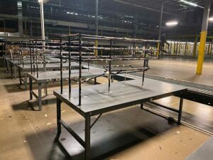 Interior of a large warehouse with several used long metal packing tables arranged in rows. The packing tables have multiple tiers of shelving above the main tabletop, providing ample storage and workspace. The area is well-lit with industrial lighting, and the concrete floor is clean. In the background, more packing tables and equipment can be seen, indicating a spacious and organized work environment.