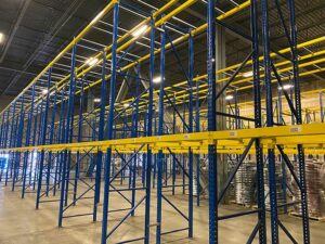 The image shows a used pallet rack system in a warehouse. The pallet racks have blue vertical upright supports and yellow horizontal beams, creating multiple levels of storage. The structure is tall and expansive, covering a large area of the warehouse. The concrete floor is clean, and the warehouse is well-lit with bright overhead lights. The racks are empty, indicating they are ready for use in storing pallets of goods. The overall setup suggests a well-organized storage solution for managing large quantities of inventory efficiently.