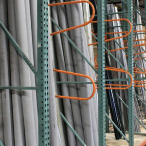 Close-up view of a warehouse storage rack holding long pipes. The pipes are stored vertically and supported by green metal frames with orange brackets. The pipes are neatly organized in rows, creating an orderly and efficient storage system. The environment appears clean and well-maintained, indicating an organized facility.
