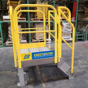 A bright yellow ErectaStep platform ladder is positioned in a warehouse. The platform has a sturdy, slip-resistant surface and is enclosed by guardrails for safety. The ErectaStep branding is prominently displayed on the side with the company’s website and contact number. The ladder is bolted to a solid, gray base for stability. In the background, there are shelves stacked with other industrial equipment and components, highlighting the organized nature of the warehouse environment.