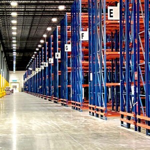 Interior of a large warehouse featuring tall rows of blue and orange metal pallet rack shelving units. The pallet rack shelves are labeled with large letters, such as "E," "F," "G," and "H," for easy identification and organization. The aisle is wide and clean, with bright industrial lighting illuminating the entire space. The high ceiling and extensive shelving create a sense of vastness and efficient storage capacity in the warehouse.