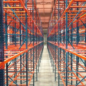 An expansive warehouse interior features numerous rows of tall, empty metal pallet rack shelving units, creating a long, symmetrical corridor. The pallet racks have bright orange horizontal supports and dark blue vertical frames, forming a striking contrast. The pallet racks extend high up towards the ceiling and deep into the distance, emphasizing the vastness and organized structure of the storage area. The concrete floor is clean and polished, reflecting the light from the high overhead industrial lighting. The overall scene highlights the extensive capacity and readiness of the warehouse for storing a large volume of inventory.