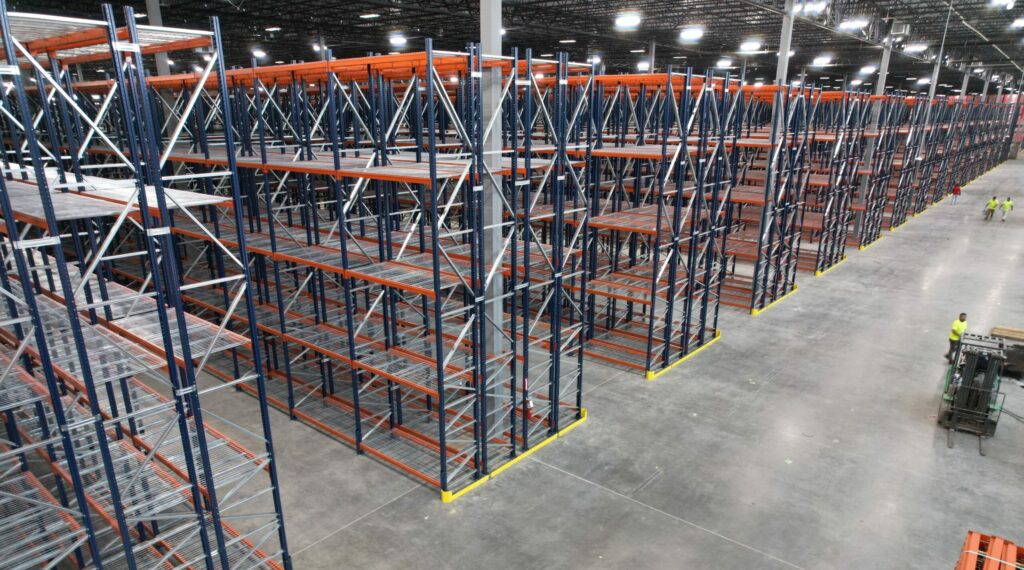 Aerial view of a large warehouse with extensive rows of tall metal pallet rack shelving units in blue and orange. The pallet rack shelves are made of beams and uprights covered in wire mesh decking, providing sturdy storage surfaces. The warehouse floor is clean and spacious, with workers visible in the distance, indicating the scale of the facility. Bright industrial lighting illuminates the entire area, highlighting the organized and expansive pallet rack storage system. The pallet rack shelving units extend far into the distance, showcasing the vast storage capacity of the warehouse.