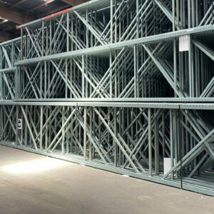 Interior of a large warehouse with stacks of green metal pallet rack shelving frames arranged in tall, neat piles. The pallet rack frames are organized along a spacious aisle, creating an orderly storage area. The high ceiling with industrial lighting illuminates the space, highlighting the extensive inventory. The concrete floor is clean, and the environment appears well-maintained and efficiently managed.