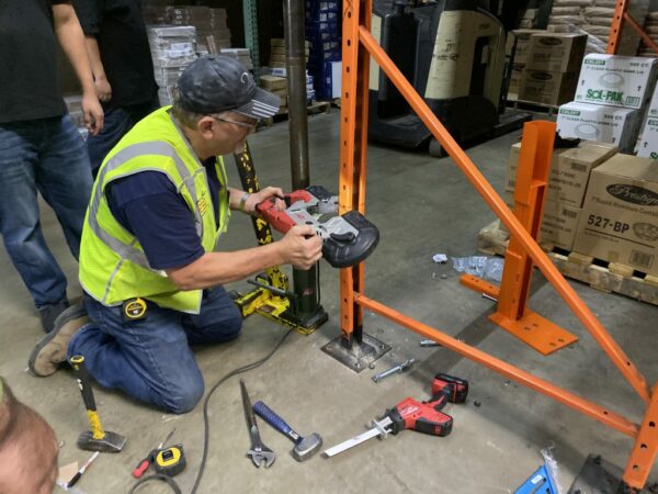 A worker wearing a yellow safety vest and a cap is kneeling on the floor of a warehouse, using a power tool to repair orange metal pallet rack upright. Various tools, including hammers, wrenches, and a measuring tape, are scattered around him. Behind the worker, there are pallets of boxes and other warehouse equipment. The scene depicts a pallet rack repair process, highlighting the hands-on work involved in maintaining warehouse infrastructure.